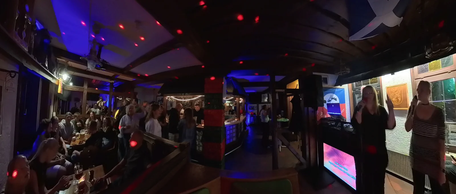 Zwei Gäste singen gemeinsam Karaoke in einer Kneipe, während weitere Gäste den Abend genießen
